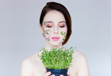 portrait of beautiful young woman with flowers on her face holding plant in pot on the wonderful grey studio backgroundの写真素材