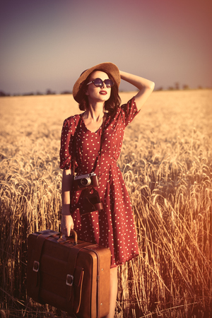 portrait of beautiful young woman with retro camera and suitcase on the wonderful field and sky backgroundの写真素材
