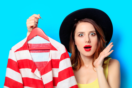 Portrait of young surprised red-haired white european woman in hat with red jacket on blue backgroundの写真素材