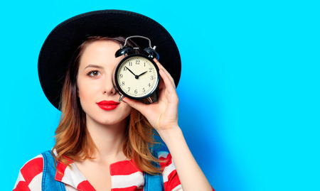 Portrait of young smiling red-haired white european woman in hat and red striped shirt with alarm...