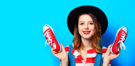 Portrait of young smiling red-haired white european woman in hat and red striped shirt with gumshoes ready for shopping on blue backgroundの写真素材