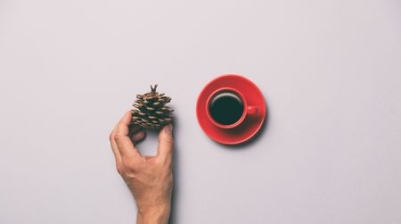 Red cup of coffee and hand holding pine cone on grey background, view aboveの写真素材