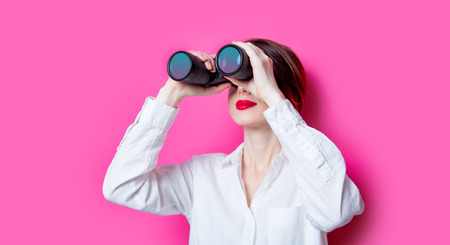 Beautiful red-haired businesswoman with binoculars on pink backgroundの写真素材