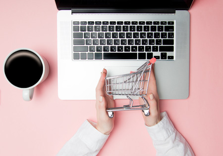 Businesswoman holding shopping cart near laptop computer on pink backgroundの写真素材