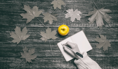 Female hand writing something in to notebook next to yellow apple and maple autumn leavesの写真素材