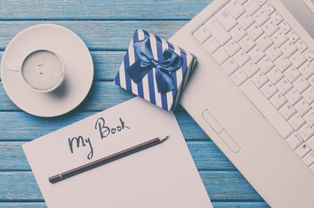a sheet of the paper, a cup of coffee, gift, pencil and laptop lying on the blue wooden tableの写真素材