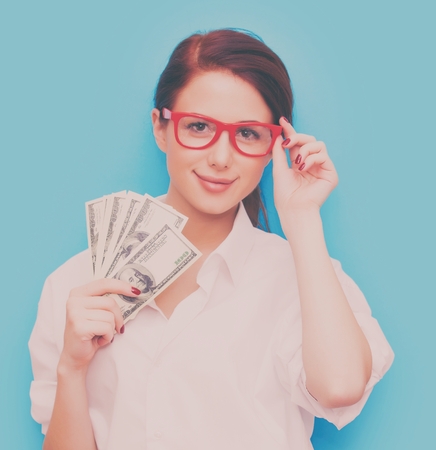 Portrait of redhead woman in red glasses with money on blue backgroundの写真素材