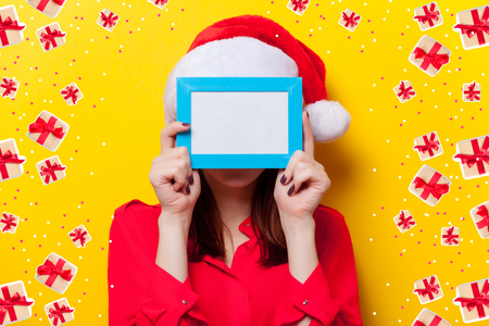 Portrait of a young redhead woman in Santa Claus hat with photo frame on yellow backgroundの写真素材