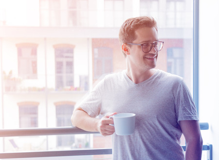 Handsome young freelancer man with cup of coffee standing near a window.の写真素材