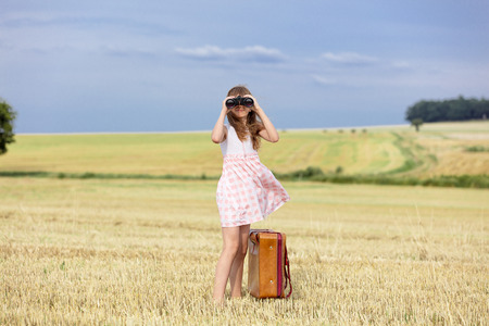 Little girl in classic dress with travel suitcase and binoculars at autumn wheat field. Tourist concept imageの写真素材