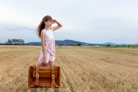 Little girl in classic dress with travel suitcase at autumn wheat field. Tourist concept imageの写真素材