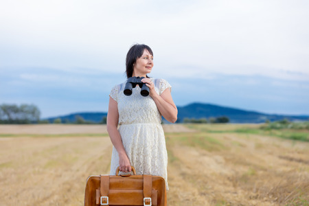 Portrait of senior woman with travel suitcase at autumn wheat field. Concept of old peope travellingの写真素材