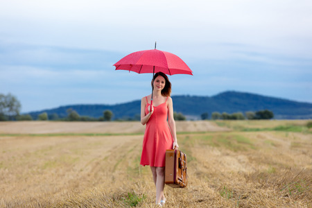 Portrait of young woman with suitcase and umbrella at autumn wheat field. Concept of old peope travelling at countrysideの写真素材
