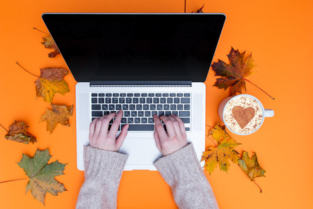 Female hands typing at laptop computer and cup of cappuccino with autumn maple leaves around at orange backgroundの写真素材