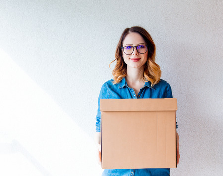 young redhead woman in jeans shirt standing on white wall with moving box. European ethnicityの写真素材