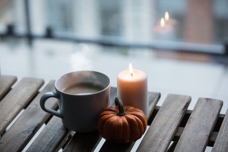 White cup of coffee or tea near a pumpkin and candle on a wooden table near a window in rainy dayの写真素材