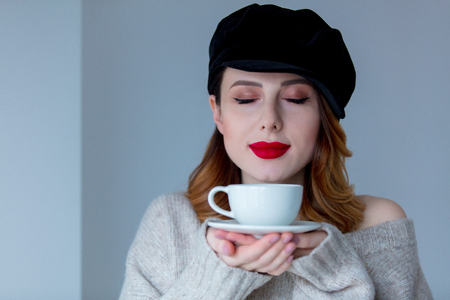 Portrait of young redhead woman in sweater and hat with cup of coffee or tea on grey background. Image made with native lightsの写真素材