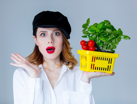 Young redhead woman in hat holding herbs of basil and red tomatoes in market basket on grey backgroundの写真素材