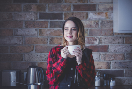 Portrait of young woman with cup of tea or coffee standing at kitchen in red shirt. Morning timeの写真素材