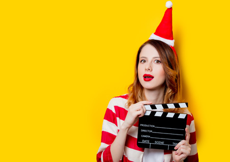 Portrait of young redhead woman in Santa Claus hat and striped shirt with filmmaking clapperboard on yellow background. Christmas timeの写真素材