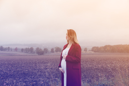 Young redhead pregnant woman in red coat standing at countryside outdoor in Autumn season timeの写真素材