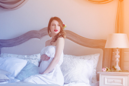 Young pregnant woman in white dress sitting in one of rooms in mansion.の写真素材