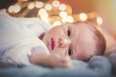 Funny little baby wearing a white clothes lying down on knitted rug in a sunny nursery.  Infant just borned, have a one week. Fairy or Christmas lights on backgroundの写真素材