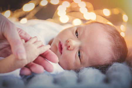 Funny little baby wearing a white clotheslying down on knitted rug in a sunny nursery.  Infant just borned, have a one week. Fairy or Christmas lights on background. Mother holding child hand.の写真素材