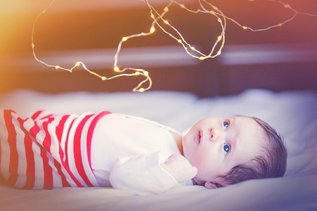 Funny little baby wearing a white clothes lying down on a bed. Infant just borned, have a one week. Fairy or Christmas lights on around.の写真素材