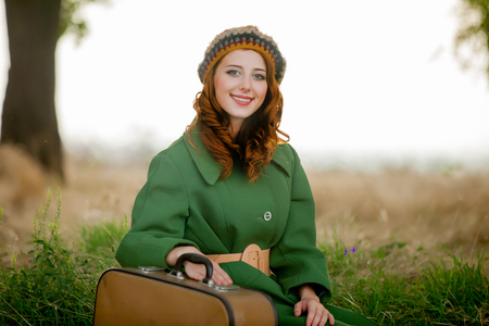 portrait of adult girl in green coat with suitcase sitting at grass at countryside outdoor.の写真素材