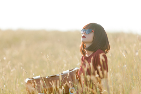 Young girl with suitcase sitting at dry grass at countrysideの写真素材