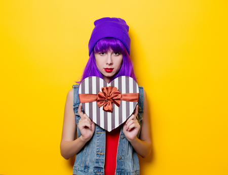 Portrait of young style hipster girl with purple hairstyle with Valentine Day gift box on yellow...