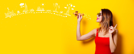 Young girl in red dress with airplane toy dreaming about world attractions on yellow backgroundの写真素材
