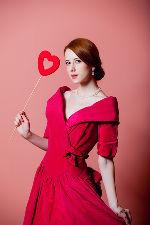 Young redhead woman in red Victorian epoch clothes with heart shape toy on pink backgroundの写真素材