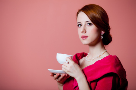 Young redhead woman in red Victorian epoch clothes with cup of tea on pink backgroundの写真素材