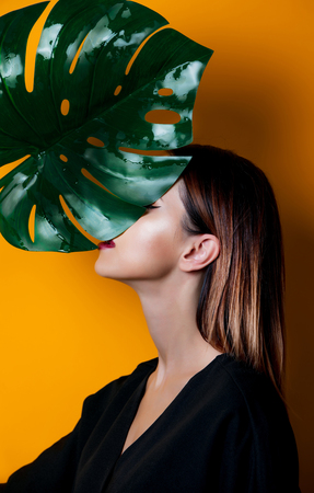 Beautiful woman in dark clothes with palm leaf on yellow color backgroundの写真素材