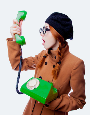 Portrait of a young redhead funny nerd girl with eyeglasses and coat talking by phone on white backgroundの写真素材