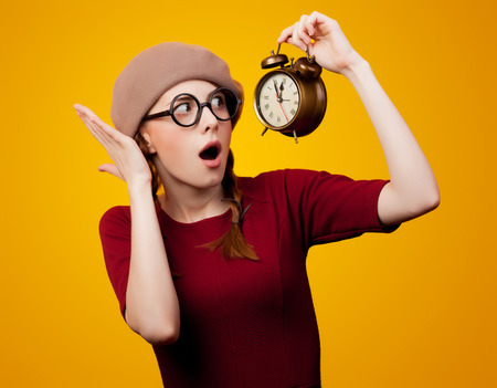 young redhead nerd girl with eyeglasses and alarm clock on yellow background