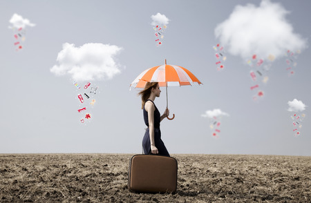Lonely girl with suitcase and umbrella at countryside field with couds and clothes rainの写真素材