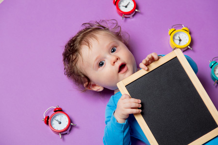 little infant baby with alarm clock and blackboard lying down on purple backgroundの写真素材