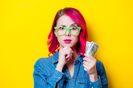 Young pink hair girl in blue shirt and green glasses holding a money. Portrait isolated on yellow backgroundの写真素材