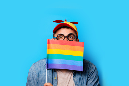 Young nerd man with noob hat holding a rainbow flag on blue backgroundの写真素材