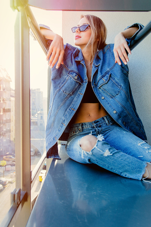 Young redhead girl in jeans clothes and sunglasses sitting on balcony alone.の写真素材