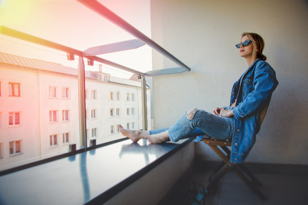 Young redhead girl in jeans clothes and sunglasses sitting on balcony alone.の写真素材