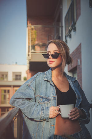 Young redhead girl in jeans clothes and cup of coffee standing on balcony alone.の写真素材