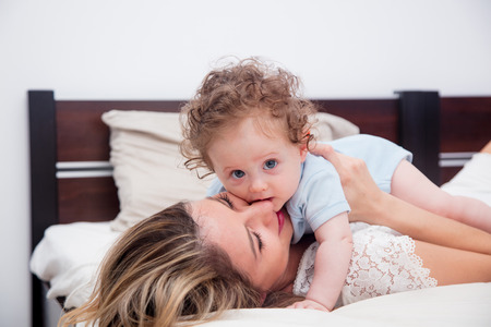 Young mother with her 7 month old little son dressed in pajamas are relaxing and playing in the bed at the weekend together, lazy morning, warm and cozy scene.の写真素材