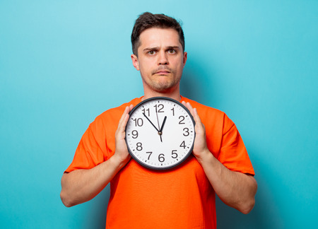 Young handsome man in orange t-shirt with big clock. Studio image on blue backgroundの写真素材