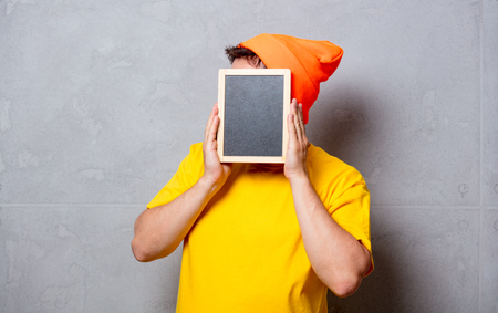 Young handsome man in yellow t-shirt and orange hat with blackboard on grey backgroundの写真素材