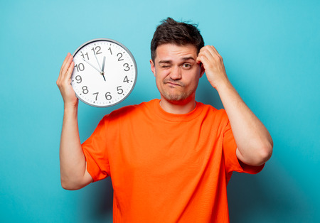 Young handsome man in orange t-shirt with big clock. Studio image on blue backgroundの写真素材