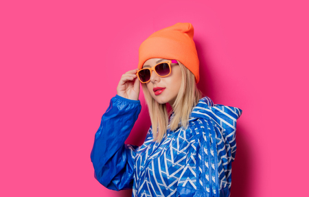 Young blonde girl in 90s sports jacket and hat with sunglasses on pink background.の写真素材
